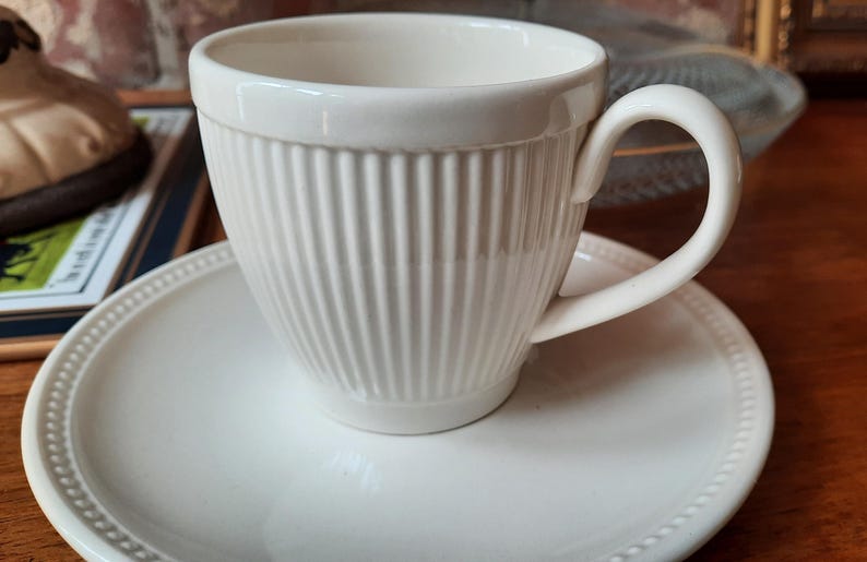 May include: A white ceramic teacup and saucer set. The teacup has vertical ribbed detailing and a curved handle. The saucer is round with a beaded edge. The set is placed on a wooden surface.
