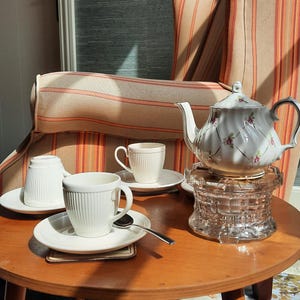 May include: A white porcelain tea set on a round wooden table. The set includes a teapot with a floral design, cups, saucers, and a glass coaster. A spoon rests on a saucer. The background features a striped armchair.