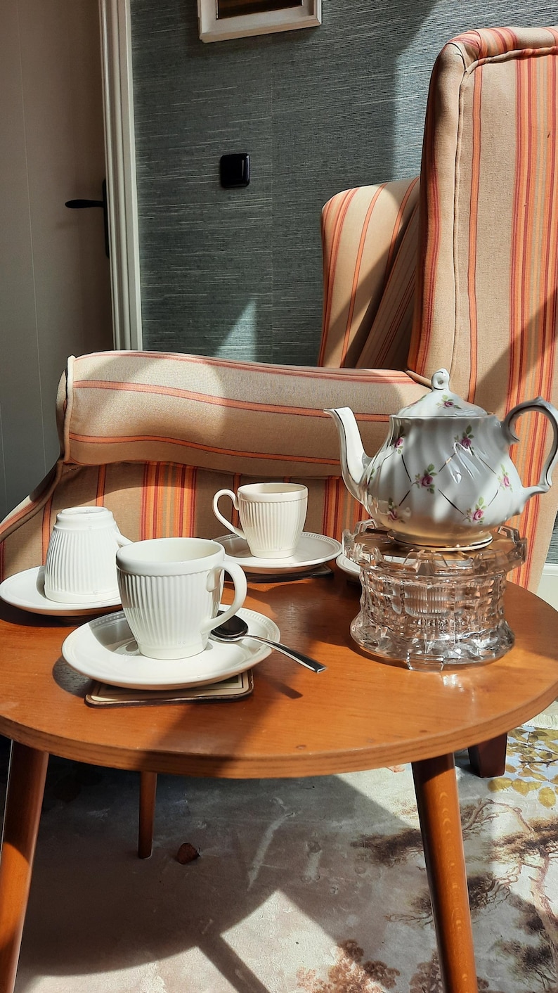 May include: A white porcelain tea set on a round wooden table. The set includes a teapot with a floral design, cups, saucers, and a glass coaster. A spoon rests on a saucer. The background features a striped armchair.