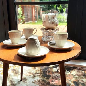 May include: A small, round wooden table with four white teacups and saucers, and a teapot. The teacups and teapot have a ribbed design. The table is set near a window with a view of a garden.