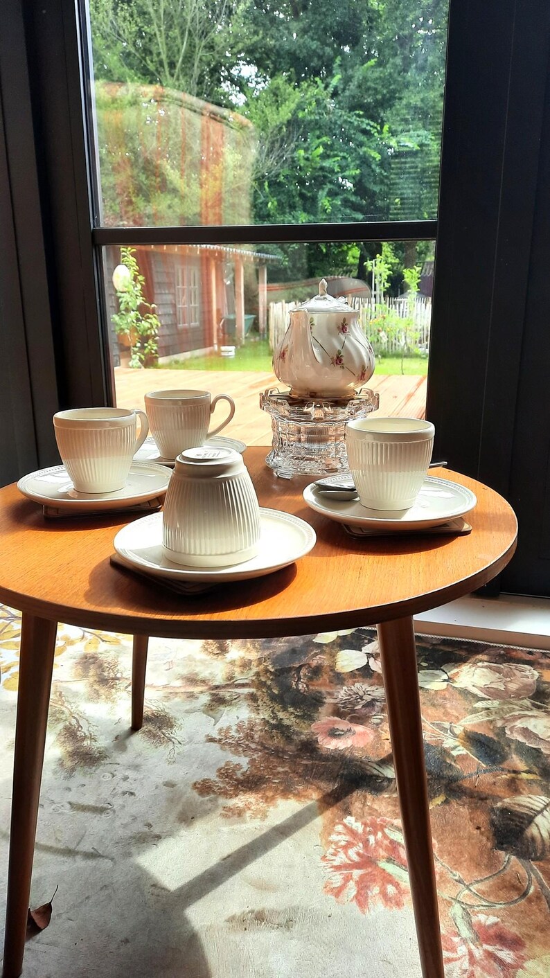 May include: A small, round wooden table with four white teacups and saucers, and a teapot. The teacups and teapot have a ribbed design. The table is set near a window with a view of a garden.
