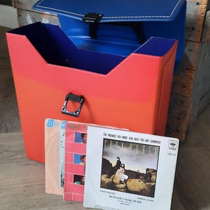 Könnte beinhalten: Eine rot-blaue Schallplatten-Aufbewahrungstasche mit schwarzem Verschluss, die mehrere Schallplatten enthält. Die Tasche ist offen und zeigt einen roten Innenraum. Einige Schallplattenhüllen sind sichtbar, darunter eine für Cock Robin.