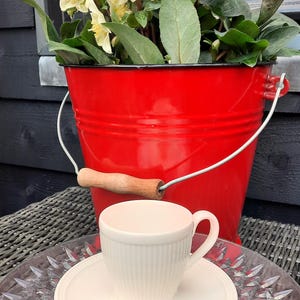 May include: A red metal bucket with a wooden handle holds a plant with yellow flowers and green leaves. A white teacup and saucer sit on a clear glass plate, resting on a woven surface.