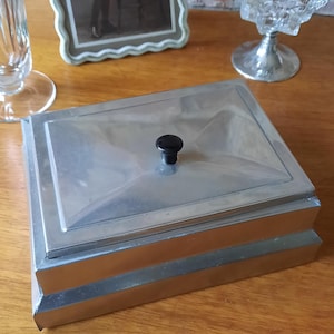 Könnte beinhalten: Eine rechteckige, silberfarbene Metallbox mit einem schwarzen Knopf auf dem Deckel. Die Box hat einen Klappdeckel und ein abgestuftes Design. Der Hintergrund umfasst ein gerahmtes Schwarz-Weiß-Foto und eine dekorative Glasschale.