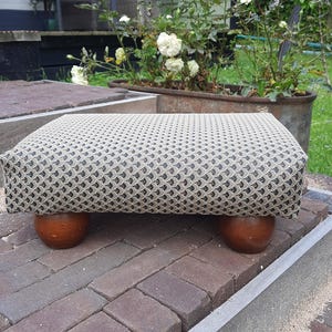 Op de afbeelding: Een rechthoekige voetenbank met een beige en zwarte stoffen bekleding met een herhalend waaiervormig patroon. De voetenbank wordt ondersteund door vier afgeronde, donkerbruine houten poten die een stabiele basis vormen voor het gestoffeerde deel.