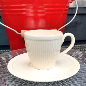 May include: A cream-colored teacup and saucer set with vertical ribbed detailing. The cup sits on a matching saucer with a raised edge. A red bucket is in the background, suggesting an outdoor setting. The teacup has a curved handle.