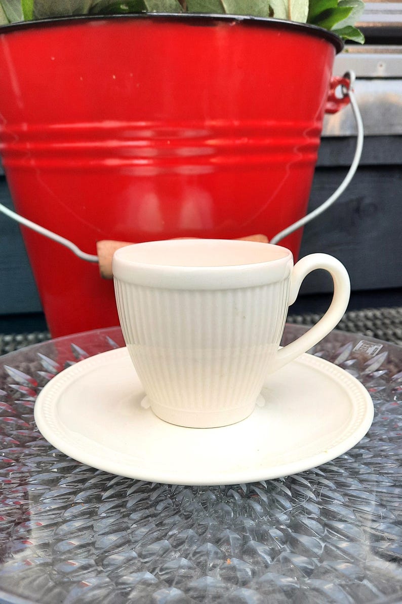 May include: A cream-colored teacup and saucer set with vertical ribbed detailing. The cup sits on a matching saucer with a raised edge. A red bucket is in the background, suggesting an outdoor setting. The teacup has a curved handle.