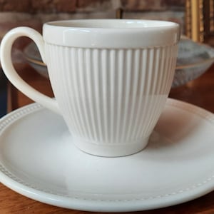 May include: A white ceramic teacup with vertical ribbed detailing sits on a matching saucer. The cup has a curved handle and a slightly flared rim. The saucer has a beaded edge. The set is on a wooden surface.