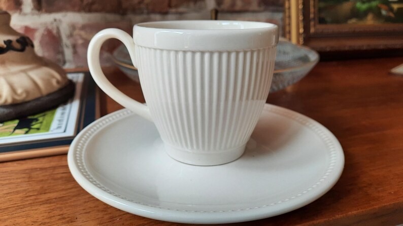 May include: A white ceramic teacup with vertical ribbed detailing sits on a matching saucer. The cup has a curved handle and a slightly flared rim. The saucer has a beaded edge. The set is on a wooden surface.