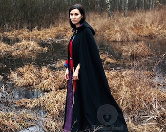 May include: A woman wearing a long, black hooded cloak stands in a marshy area. The water is dark and the ground is covered in dry, brown reeds.