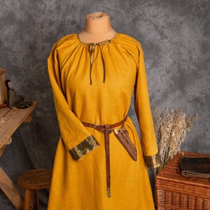May include: A long, yellow linen dress with a gathered neckline and long sleeves. The dress has a brown leather belt and a green and gold trim around the neckline and sleeves.
