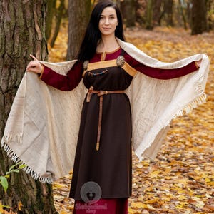 May include: A medieval-style dress in shades of brown and burgundy, with a cream-colored shawl. The dress features a brown leather belt and decorative clasps. The background shows a forest with fallen leaves.