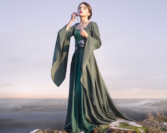 May include: A woman in a long, green dress stands on a rock overlooking a misty landscape. She holds a small, silver pendant on a chain.