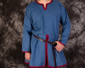 Op de afbeelding: Een man draagt ​​een blauwe tuniek met rode bies en een bruine leren riem. De tuniek heeft een V-hals en lange mouwen. De man staat tegen een grijze, getextureerde achtergrond.