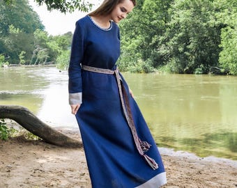 May include: A long, flowing, navy blue dress with long sleeves and a decorative belt. The dress has a white trim at the hem and cuffs. The model is standing on a sandy beach near a river.