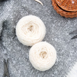 May include: Two balls of white yarn, a pair of scissors, a wooden yarn needle, and a wicker basket with a lid on a grey and white patterned fabric.