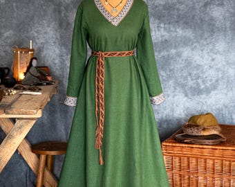 May include: A long, green dress with long sleeves and a V-neck, displayed on a mannequin. The dress features white and beige trim along the neckline, cuffs, and hem. A brown woven belt cinches the waist. A necklace is visible.