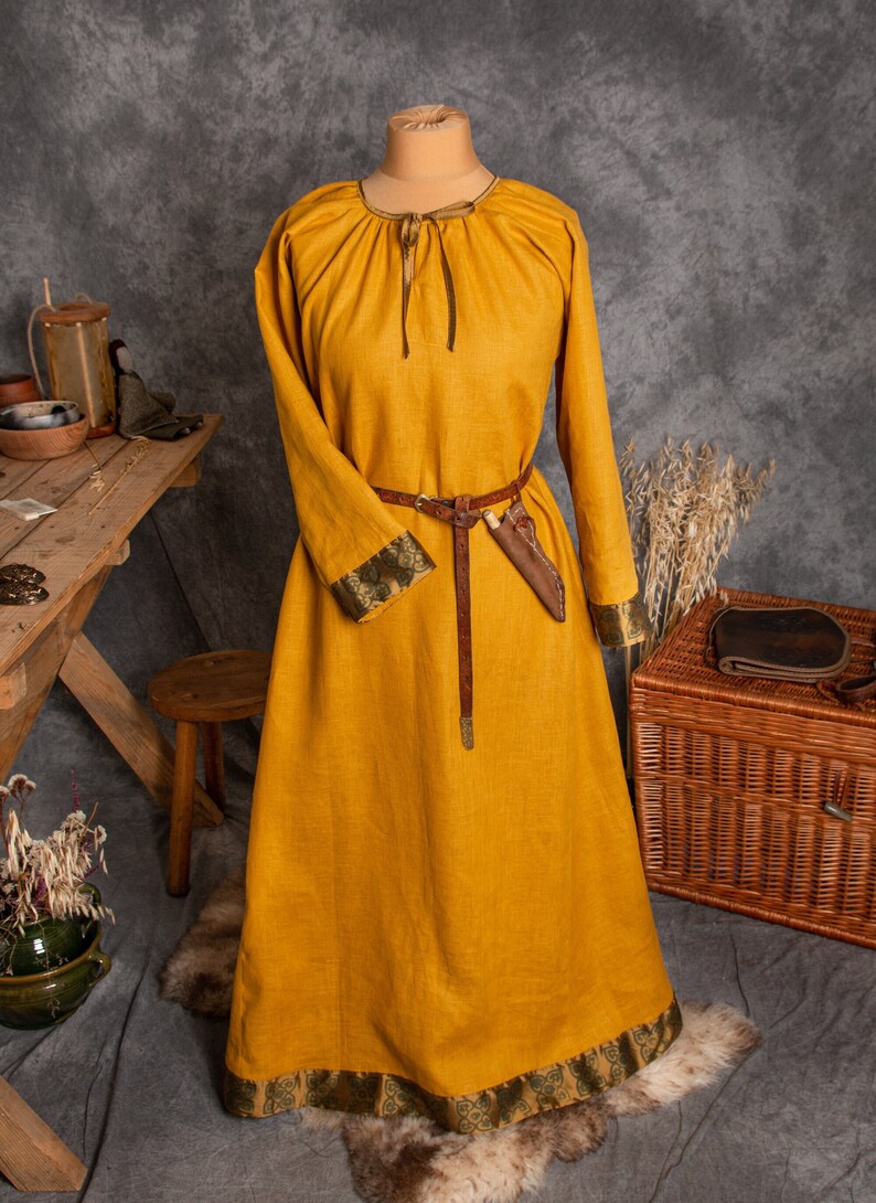 May include: A long, yellow linen dress with a gathered neckline and long sleeves. The dress has a brown leather belt and a green and gold trim around the neckline and sleeves.