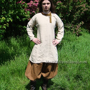 May include: A person wearing a long-sleeved, light-colored shirt with a brown belt and brown pants. The shirt has a decorative trim around the neckline. The person is standing in a grassy field.