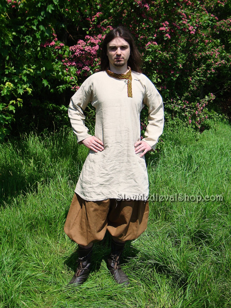 May include: A person wearing a long-sleeved, light-colored shirt with a brown belt and brown pants. The shirt has a decorative trim around the neckline. The person is standing in a grassy field.