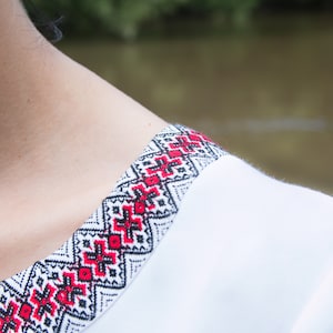 Op de afbeelding: Wit geborduurd shirt met een rood en zwart geometrisch patroon rond de halslijn.