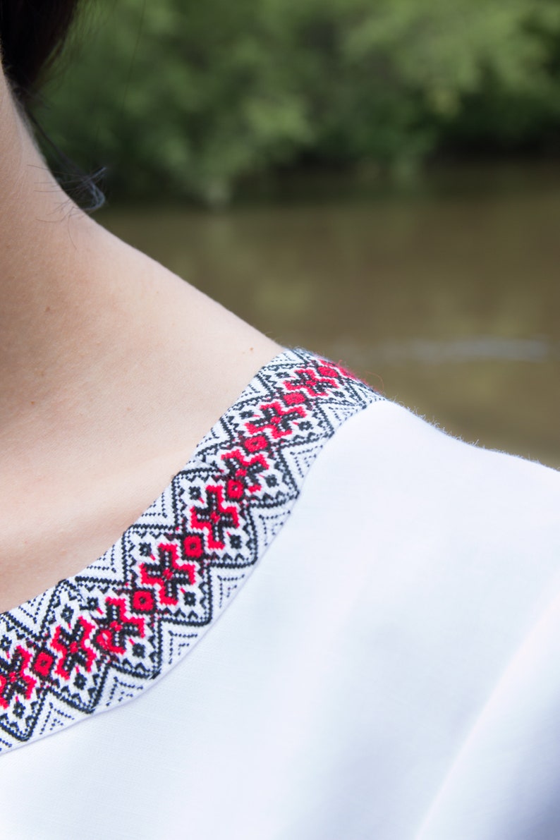 Op de afbeelding: Wit geborduurd shirt met een rood en zwart geometrisch patroon rond de halslijn.