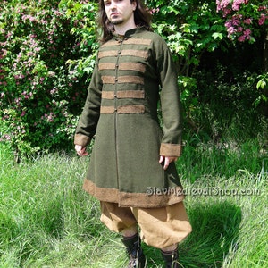 May include: A long, dark green coat with brown trim and a high collar. The coat has a button closure and is worn over brown pants. The person is standing in a grassy field.