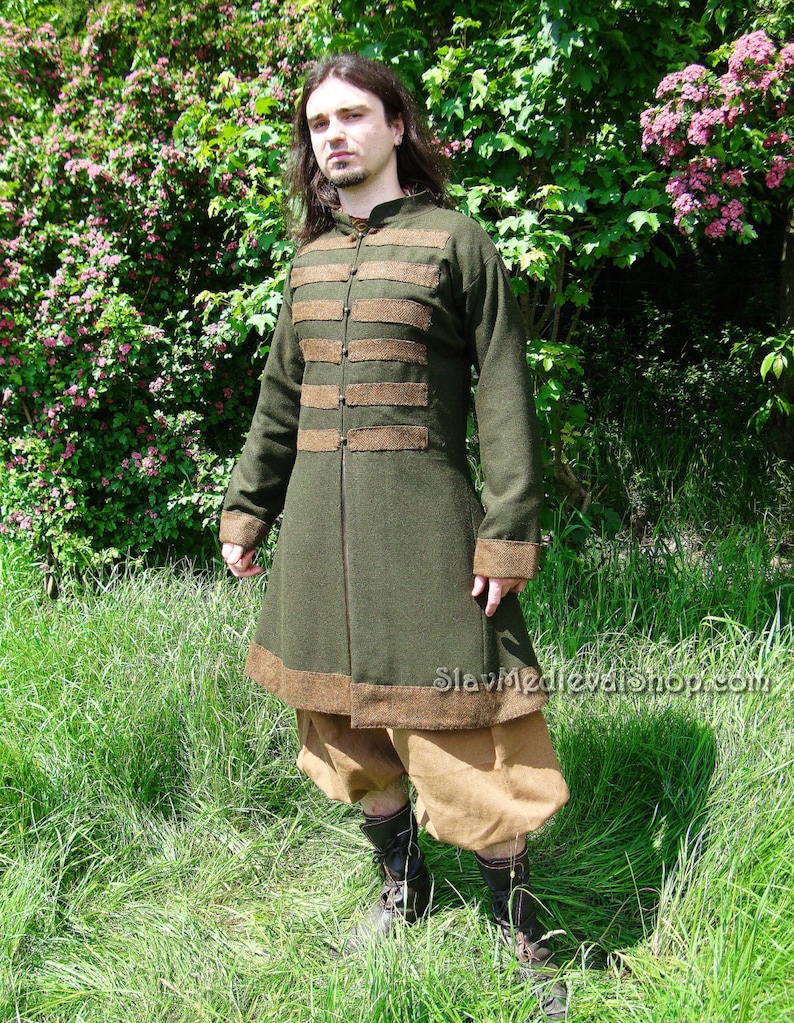 May include: A long, dark green coat with brown trim and a high collar. The coat has a button closure and is worn over brown pants. The person is standing in a grassy field.