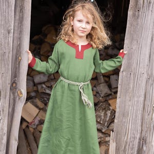 Puede incluir: Un vestido túnica verde de manga larga y hasta el suelo, con un ribete de cuello en rojo oscuro y puños a juego. La prenda se ajusta a la cintura con un cinturón fino de cordón trenzado en color blanco roto y verde, atado con un nudo.