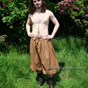 May include: A person wearing brown linen pants with a gathered waist and brown leather boots. The pants are loose fitting and have a wide leg. The person is standing in a field of green grass.