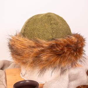 Può includere: Un cappello di lana con motivo verde e marrone con un bordo in pelliccia sintetica. Il cappello è su una superficie di legno.