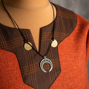 May include: A brown and orange tunic with a black leather cord necklace featuring a silver crescent moon pendant and two gold coin charms.