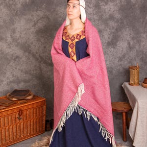 K&ouml;nnte beinhalten: Eine Frau in einem mittelalterlichen blauen Kleid mit goldenen Verzierungen und einem rosa gewebten Schal mit Fransen. Das Kleid hat einen dekorativen Ausschnitt. Die Frau tr&auml;gt eine wei&szlig;e Kopfbedeckung und ein d&uuml;nnes Stirnband. Eine Holzkiste und ein Hocker sind im Hintergrund.