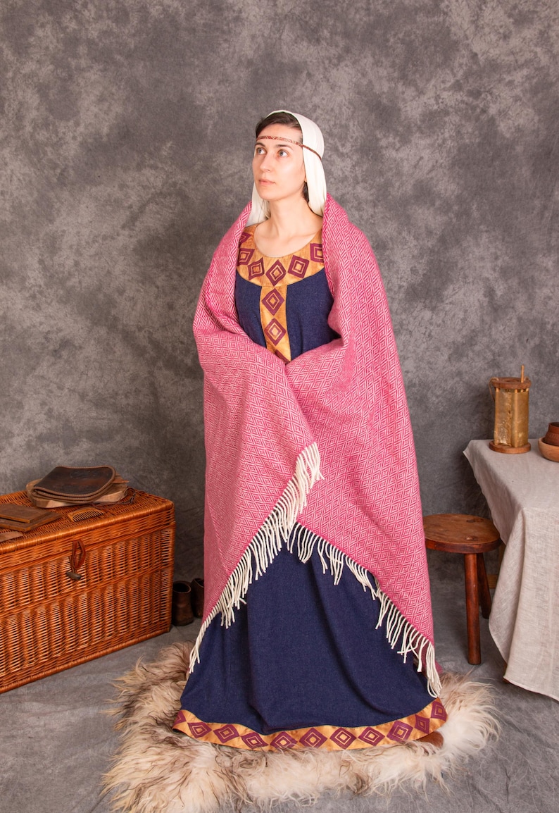K&ouml;nnte beinhalten: Eine Frau in einem mittelalterlichen blauen Kleid mit goldenen Verzierungen und einem rosa gewebten Schal mit Fransen. Das Kleid hat einen dekorativen Ausschnitt. Die Frau tr&auml;gt eine wei&szlig;e Kopfbedeckung und ein d&uuml;nnes Stirnband. Eine Holzkiste und ein Hocker sind im Hintergrund.