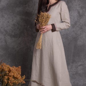 May include: A long, beige linen dress with long sleeves and a V-neckline. The dress features brown trim around the neckline and cuffs, with red stitching. The woman is holding a bundle of dried flowers. The dress is a medieval or renaissance style.