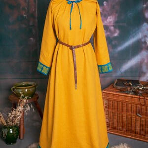 May include: A long, yellow linen dress with blue and green trim. The dress has a gathered neckline and a brown leather belt. The dress is displayed on a mannequin against a rustic background.