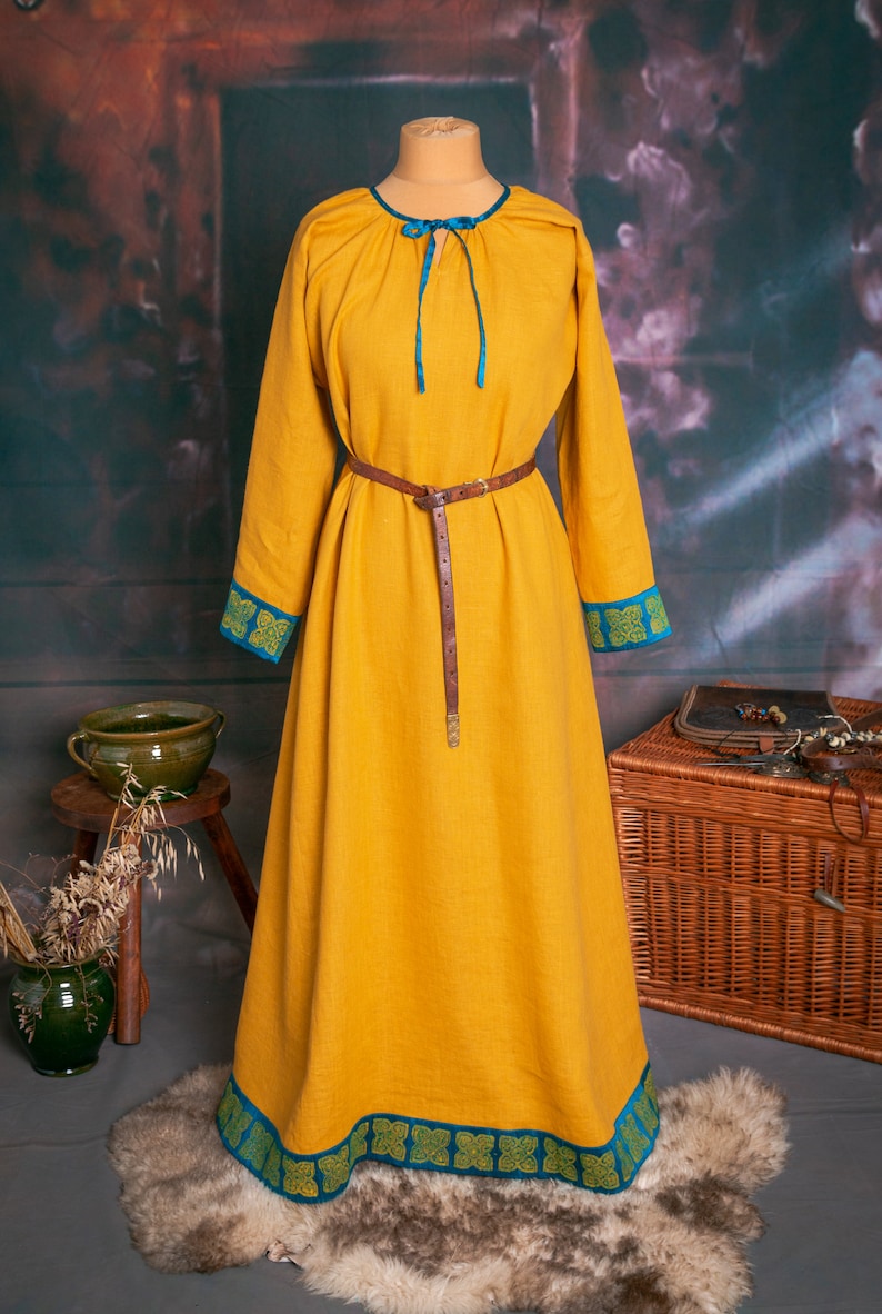 May include: A long, yellow linen dress with blue and green trim. The dress has a gathered neckline and a brown leather belt. The dress is displayed on a mannequin against a rustic background.