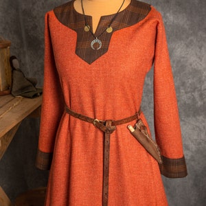 May include: A long, orange dress with brown trim and a leather belt. The dress has a V-neck and is made of a textured fabric. The belt has a metal buckle and a small leather pouch attached to it.