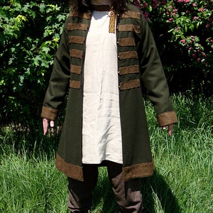 May include: A long, dark green coat with brown trim and a white shirt underneath. The coat has several brown bands around the waist and sleeves. The person is standing in a grassy field.