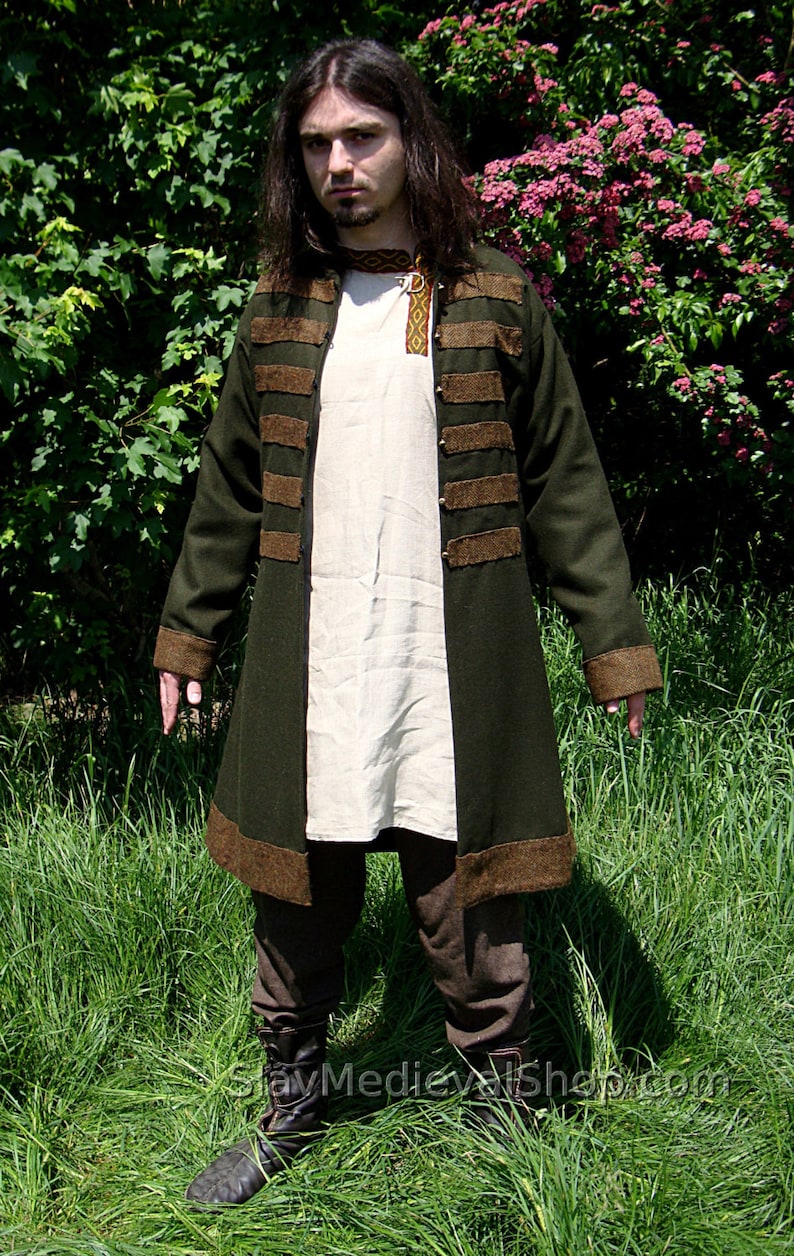 May include: A long, dark green coat with brown trim and a white shirt underneath. The coat has several brown bands around the waist and sleeves. The person is standing in a grassy field.