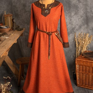 May include: A long, orange dress with brown trim and a brown leather belt. The dress is made of a textured fabric and has a simple, elegant design.