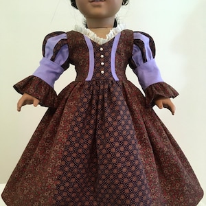 Puede incluir: Una mu&ntilde;eca con un vestido burdeos con estampado floral y mangas y detalles en lavanda. El vestido tiene un cuello de encaje blanco y peque&ntilde;os botones de perlas en la parte delantera. La falda es amplia y fruncida.