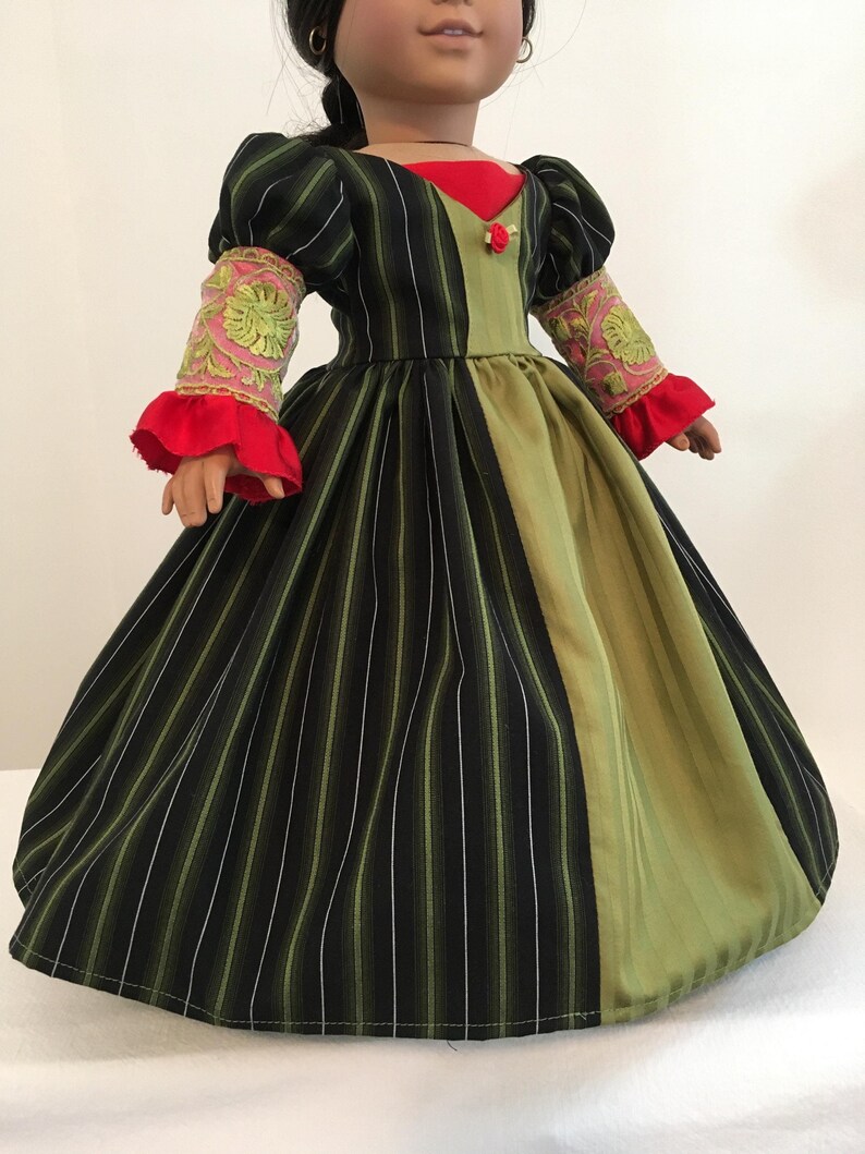 May include: A doll wearing a striped black and green dress with a red bodice and sleeves. The sleeves have pink and gold embroidered details and red ruffles. A small red rose is on the bodice.