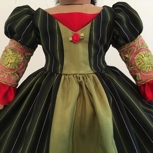 May include: A doll-sized dress with a black and green striped bodice and skirt. The dress features a red neckline and cuffs, with pink and gold embroidered sleeves. A small red rose adorns the bodice, creating a vintage aesthetic.