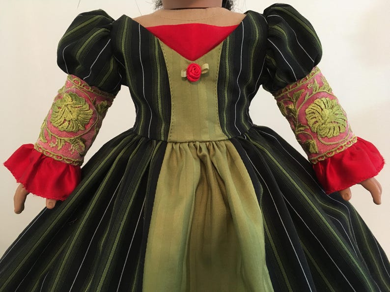 May include: A doll-sized dress with a black and green striped bodice and skirt. The dress features a red neckline and cuffs, with pink and gold embroidered sleeves. A small red rose adorns the bodice, creating a vintage aesthetic.
