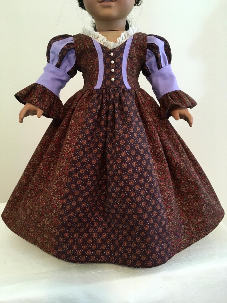 Puede incluir: Una mu&ntilde;eca con un vestido burdeos con estampado floral y mangas y detalles en lavanda. El vestido tiene un cuello de encaje blanco y peque&ntilde;os botones de perlas en la parte delantera. La falda es amplia y fruncida.