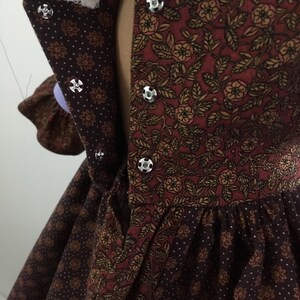 Puede incluir: Primer plano de un vestido de estilo vintage con un estampado floral en tonos burdeos, marr&oacute;n y dorado. El vestido presenta un cuello alto con botones plateados y un ribete de encaje. La tela tiene un estampado de lunares en la falda.
