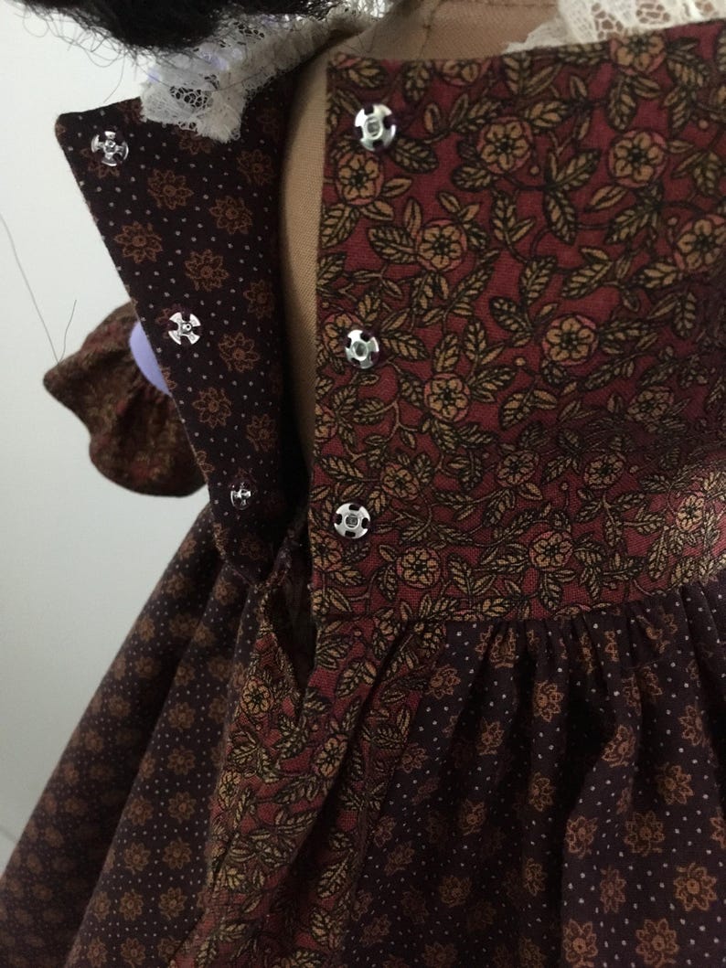 Puede incluir: Primer plano de un vestido de estilo vintage con un estampado floral en tonos burdeos, marr&oacute;n y dorado. El vestido presenta un cuello alto con botones plateados y un ribete de encaje. La tela tiene un estampado de lunares en la falda.