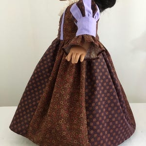 Puede incluir: Una mu&ntilde;eca con un vestido estampado marr&oacute;n y morado. El vestido presenta un cuello alto con ribete de encaje blanco y las mangas est&aacute;n acentuadas con tela lavanda. La falda es amplia y tiene un estampado floral y de lunares.