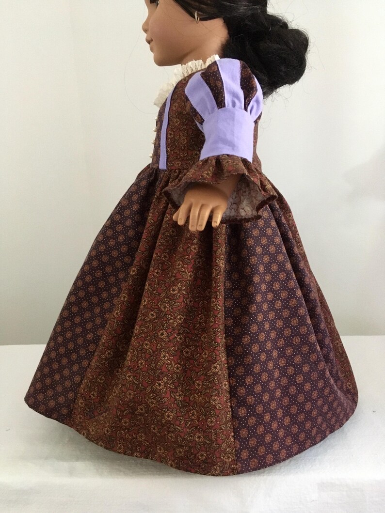 Puede incluir: Una mu&ntilde;eca con un vestido estampado marr&oacute;n y morado. El vestido presenta un cuello alto con ribete de encaje blanco y las mangas est&aacute;n acentuadas con tela lavanda. La falda es amplia y tiene un estampado floral y de lunares.